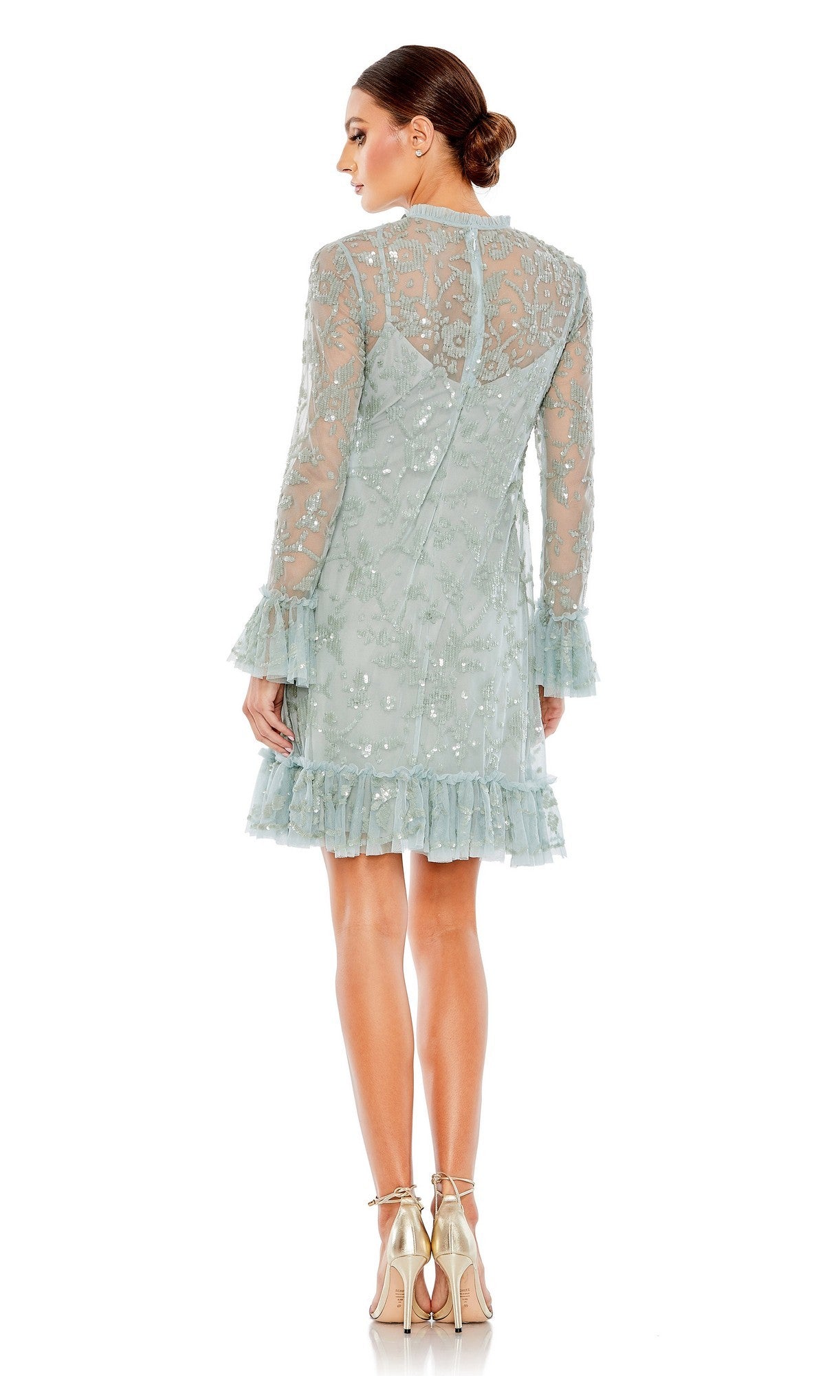A woman stands in profile against a white background, wearing the Mac Duggal 9139 short party dress—a light blue, long-sleeve, knee-length lace dress with ruffle hem and cuffs, delicate sequin detailing—and gold high-heeled shoes.